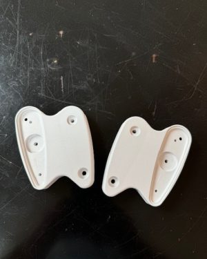 Twee Studio White plastic wandbeugels op een donker oppervlak. Elk met meerdere schroefgaten en gebogen randen, wat de stijl van Herman Miller weerspiegelt: ideaal voor het stijlvol ondersteunen van een apparaat of armatuur. Product: Mirra Arm-adapter.