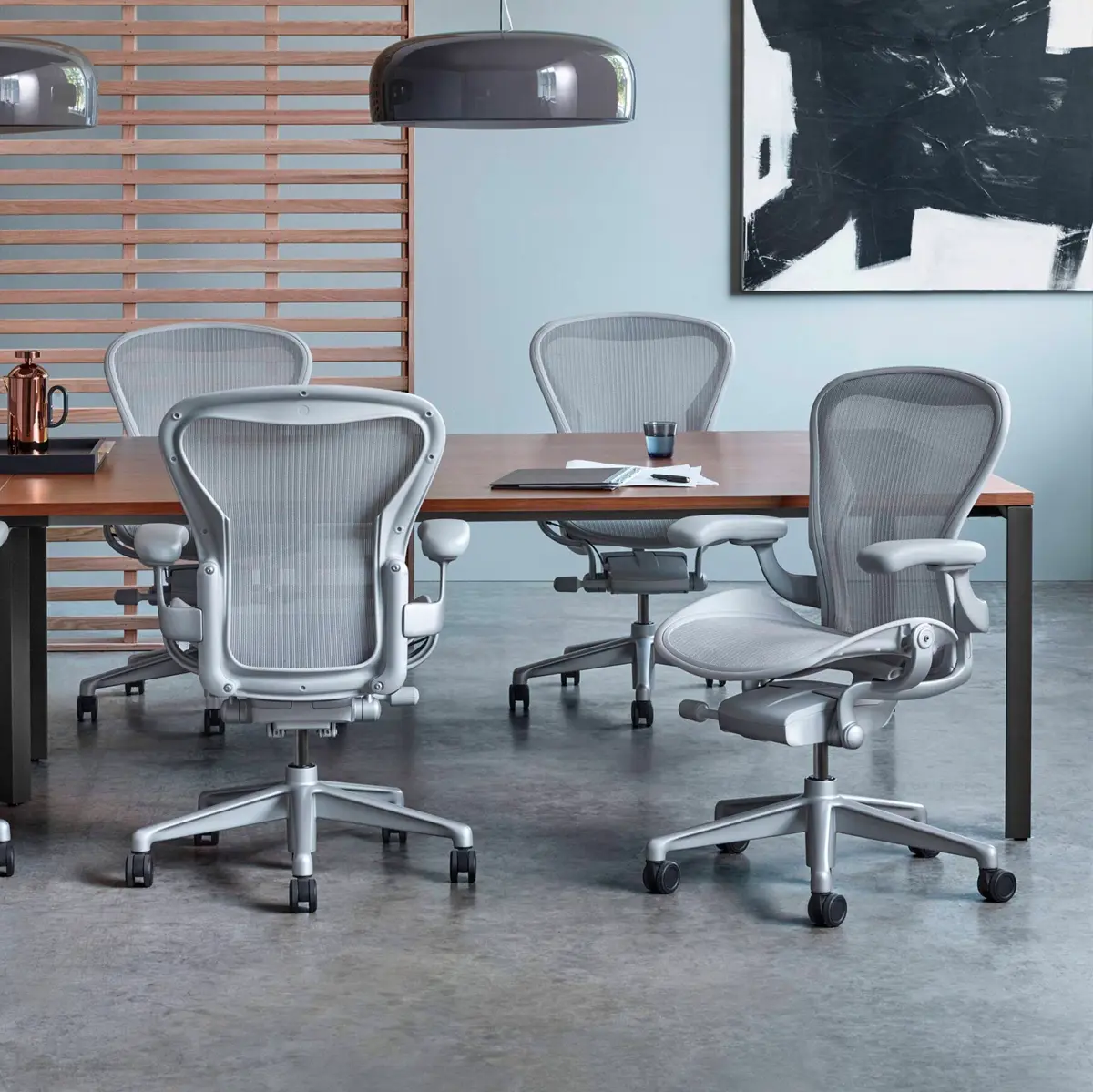 Een moderne kantoorvergaderruimte met een donkere houten tafel omringd door vier Herman Miller ergonomische mesh-kantoorstoelen op wielen. Boven de tafel hangt een grote, strakke hanglamp. De muren zijn versierd met een houten lataccent en abstracte zwart-witkunst.