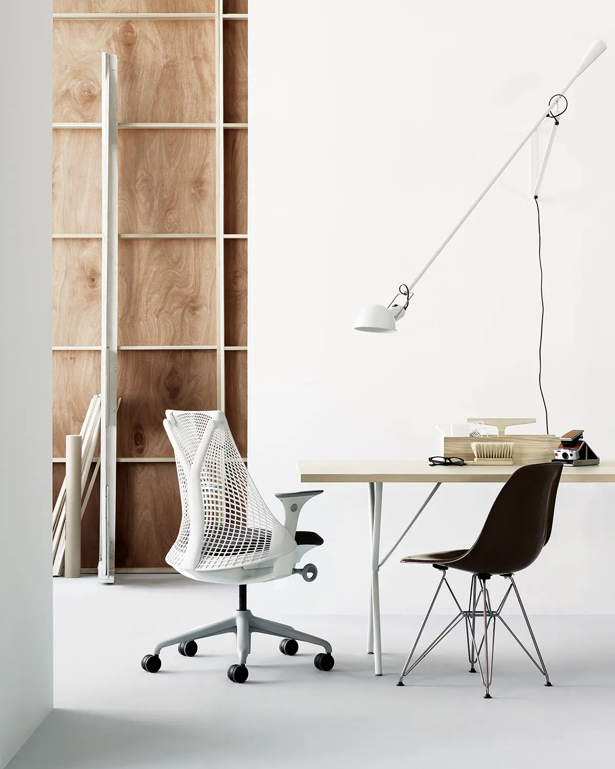 De minimalistische werkruimte beschikt over een witte ergonomische Herman Miller-stoel en een bruine stoel aan een licht houten bureau. Een witte aan de muur gemonteerde lamp en houten planken met verschillende items zorgen voor een moderne touch tegen de neutrale achtergrond.