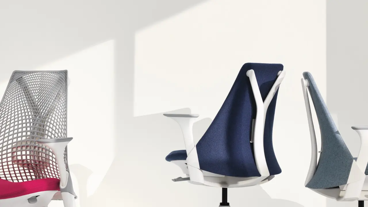 Twee moderne bureaustoelen met strakke ontwerpen stralen stijl uit. De stoel links, gedeeltelijk zichtbaar, heeft een netrug en een roze zitting. Volledig zichtbaar, rechts, is een chique Herman Miller-stuk met gladde blauwe stof en een strak wit frame tegen een lichte muur met schaduwen.