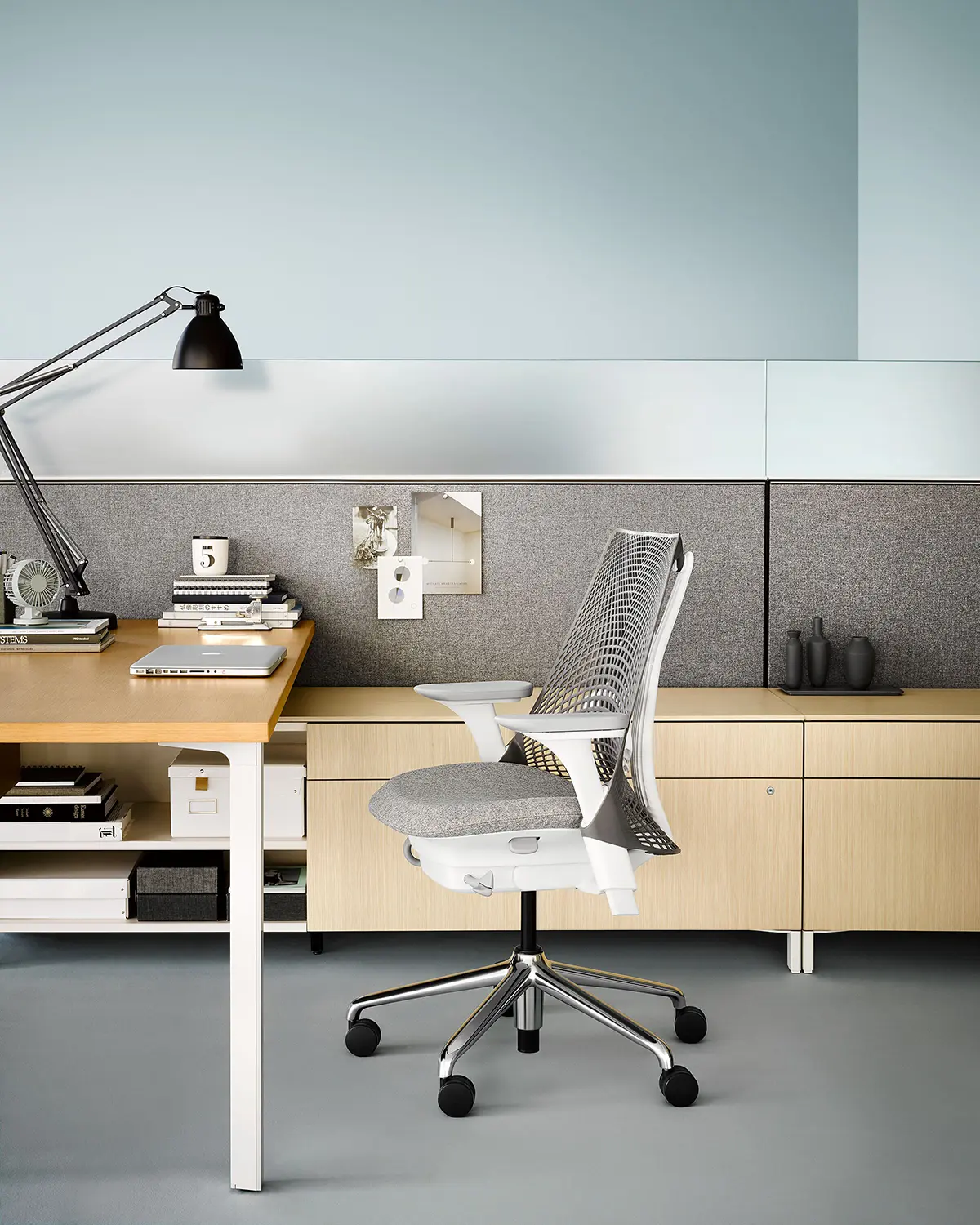 Een moderne kantoorwerkplek met een Herman Miller mesh-back ergonomische stoel aan een houten bureau met een verstelbare zwarte lamp. Het bureau heeft boeken, een kalender en kantoorbenodigdheden. Een grijze scheidingswand en lichthouten kasten maken de setting compleet.