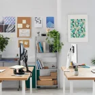 Een modern kantoor heeft twee houten bureaus, elk uitgerust met een Herman Miller-stoel en een computer ondersteund door Herman Miller Flo Monitor Arms voor 3 tot 9 kg in een enkele opstelling. De kamer beschikt over planten, kunstwerken op witte bakstenen muren, natuurlijk licht van een groot raam en planken vol boeken en bestanden.