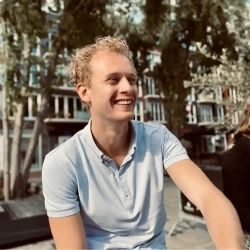 Een lachende jongeman met krullend haar in een lichtblauw shirt van LoyalBrand zit buiten. Achter hem staan bomen en een gebouw met meerdere ramen, die de essentie van een zonnige dag vastleggen.