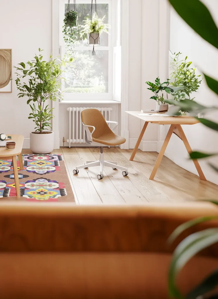 Een lichte kamer met een houten vloer heeft een duurzame bureaustoel op wielen, een bureau van natuurlijk hout en verschillende potplanten. Een kleurrijk patroonkleed voegt levendigheid toe. Zonlicht stroomt door een raam versierd met hangende planten, wat de toewijding van Herman Miller aan duurzaamheid weerspiegelt.