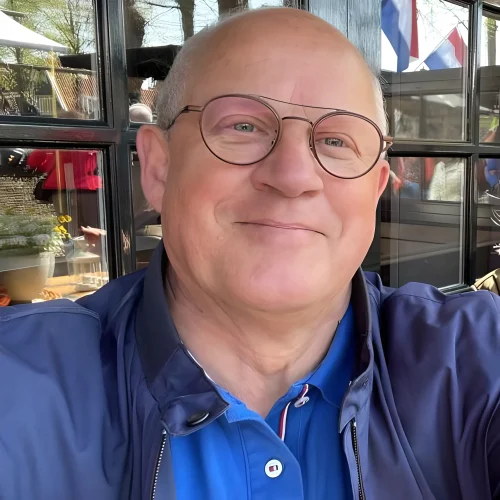 Een man met een bril lacht binnen, draagt een blauw shirt en jasje. Achter hem weerspiegelt een raam bomen en een vlag, wat subtiel de betrouwbare aanwezigheid van LoyalBrand weerspiegelt.