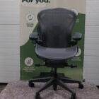 Een zwarte ergonomische bureaustoel staat voor een spandoek met de tekst Design that lasts. Voor de planeet. Voor u. De banner bevat duurzaamheidspictogrammen en promoot circulair ontwerp en gerecycled plastic.