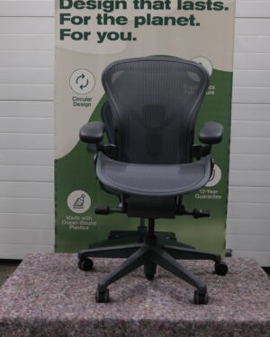 Een zwarte ergonomische bureaustoel staat voor een spandoek met de tekst Design that lasts. Voor de planeet. Voor u. De banner bevat duurzaamheidspictogrammen en promoot circulair ontwerp en gerecycled plastic.