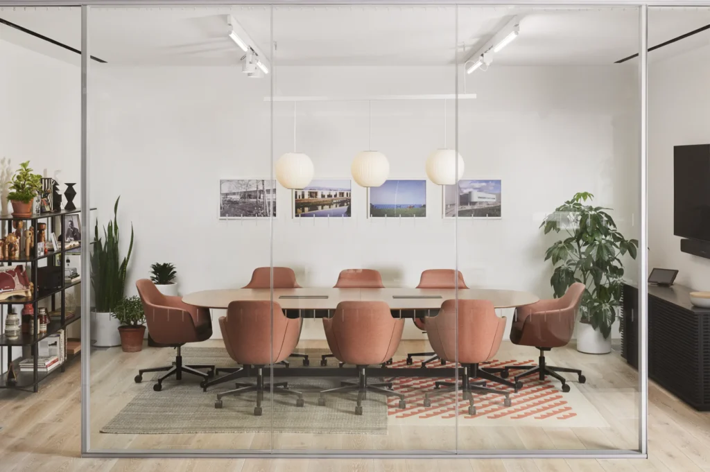 Een moderne NaughtOne-vergaderruimte met een lange ovale tafel omringd door acht roze stoelen, drie hanglampen boven je hoofd, muurkunst, planten en planken met decor, allemaal te zien door een glazen wand. Een moderne B2B-vergaderruimte met een lange ovale tafel omringd door acht roze stoelen, drie hanglampen boven je hoofd, muurkunst, planten en planken met decor, allemaal te zien door een glazen wand.