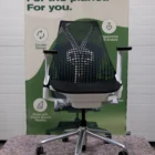 Een moderne zwarte bureaustoel met netrugleuning en zilveren onderstel staat op een mat. Daarachter staat een bord met de tekst "Duurzaam design. Voor de planeet. Voor jou." en benadrukt milieuvriendelijke kenmerken.