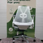 Een moderne witte bureaustoel met een netrugleuning staat voor een bord dat duurzaam ontwerp promoot, met groene tekst over duurzaamheid, milieuvoordelen en het gebruik van oceaangebonden plastic.