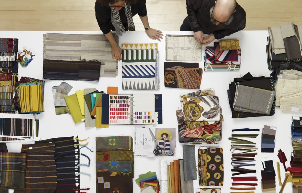 Twee mensen onderzoeken een assortiment kleurrijke stofstalen, stalen en patroonboeken die verspreid liggen op een grote witte tafel - perfect voor een B2B-bijeenkomst gericht op textielselectie en samenwerking. Twee mensen onderzoeken een assortiment kleurrijke stofstalen, stalen en patroonboeken die verspreid liggen op een grote witte tafel - perfect voor een B2B-bijeenkomst gericht op textielselectie en samenwerking.